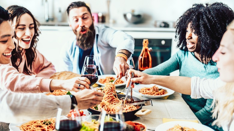 A group of joyful people sitting at the table, taking pasta from the pan at the center