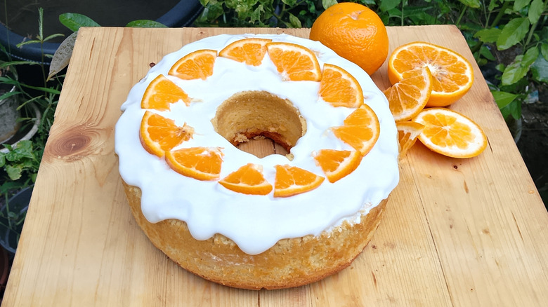 A glazed orange soda cake topped with oranges next to sliced oranges on a wooden table
