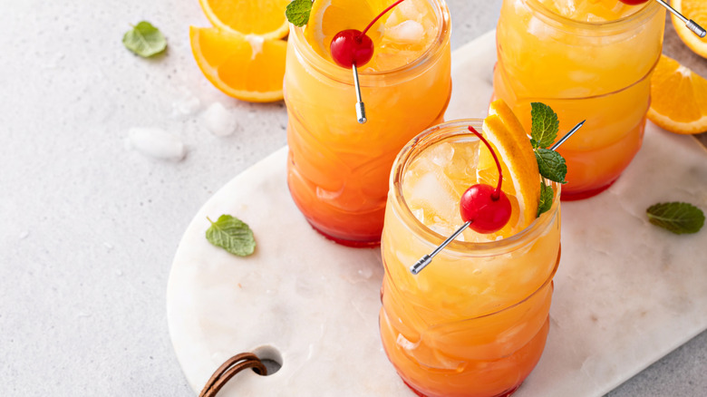 Three pineapple orange cocktails in decorative glasses, garnished with cherry, mint, and orange