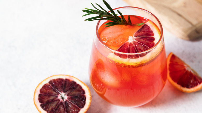 A glass with a blood orange soda cocktail garnished with an orange slice and rosemary