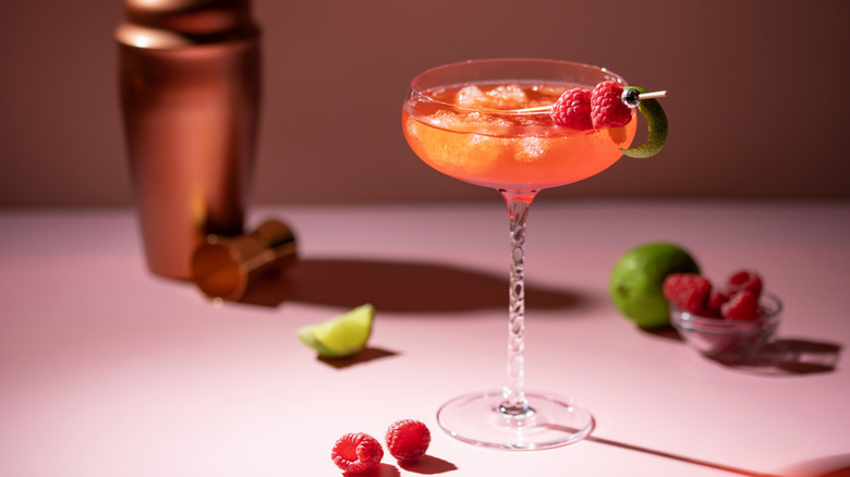 A coupe glass containing a red cocktail garnished with raspberries and a lime twist