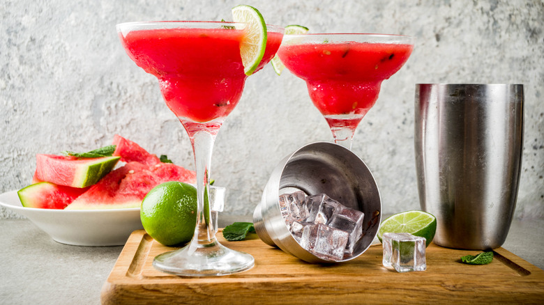 Watermelon cocktails in martini glasses, on a table next to limes, slices of watermelon, and a cocktail shaker