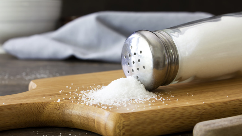 A salt shaker resting on its side on a wooden cutting board with a pile of salt in front of it