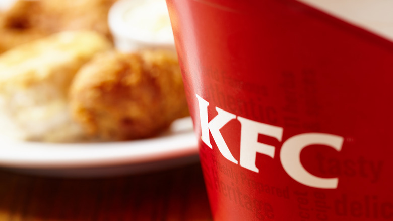 a close-up photo of a KFC bucket with a plate of chicken and biscuits blurry in the background
