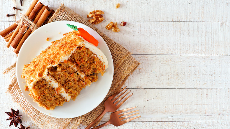 Carrot cake slice with frosting on a white plate with forks, spices, and nuts
