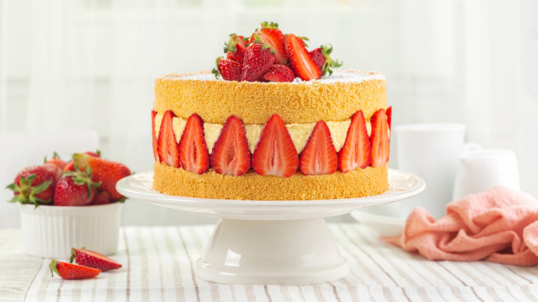 Strawberry genoise cake on a white cake stand next to a bowl of strawberries