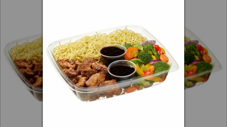 Kirkland signature yakisoba chicken stir fry kit website photo with noodles, chicken, two plastic containers of sauce, assorted vegetables all in a rectangular plastic container against a white background.