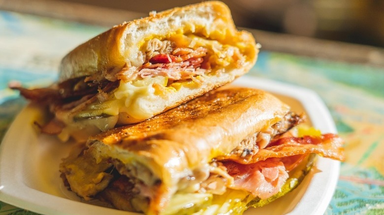 Sandwich sliced in half, stuffed with thinly sliced ham, pulled pork, cheese, and pickles on a white plate