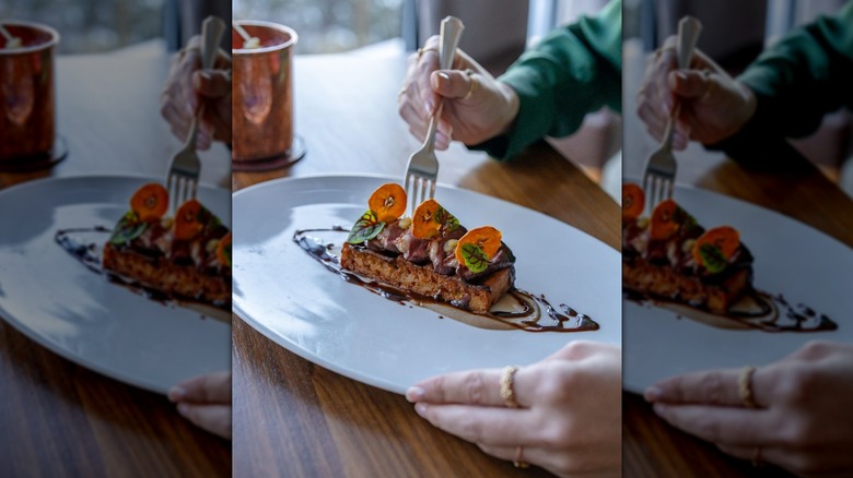 Hand holding a fork digging into a cooked duck with bananas, waffles, bright orange and green leaf garnishes, and a chocolate drizzle on a white plate