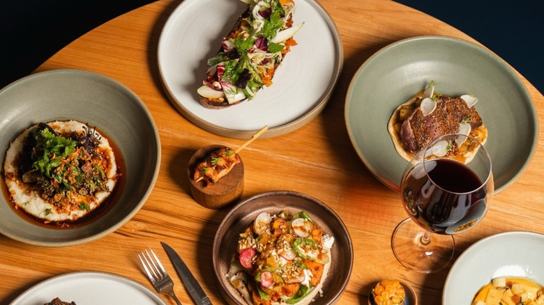 Assorted plates of delicate, sophisticated dishes scattered on wooden table, served on green and white ceramic plates, surrounded by cutlery and a single glass of red wine