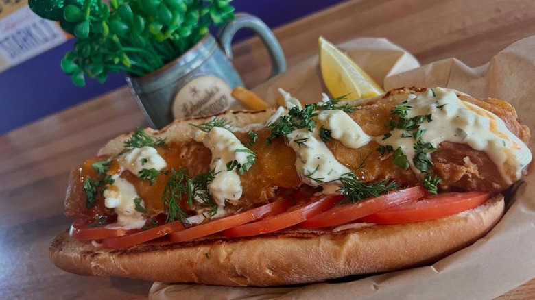 Fried cod "hot dog" stuffed in a hot dog bun lined with tomato slices and topped with tartar sauce and dill, served with a lemon slice