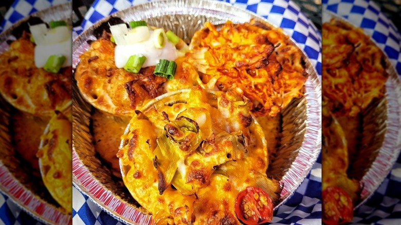 Three small containers of different types of mac and cheese and various toppings, all crispy on top, sitting in an aluminum foil plate on blue-checkered paper