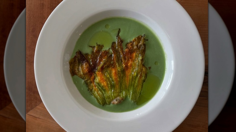 Verdant green soup in a shallow white bowl with a purple and orange fried squash blossom floating on top