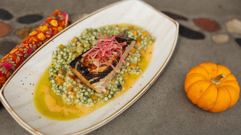 Piece of fish with charred top served in a white, boat-style plate with a yellow-green sauce, some kind of peas floating in it, and a pile of pink strips of something for garnish