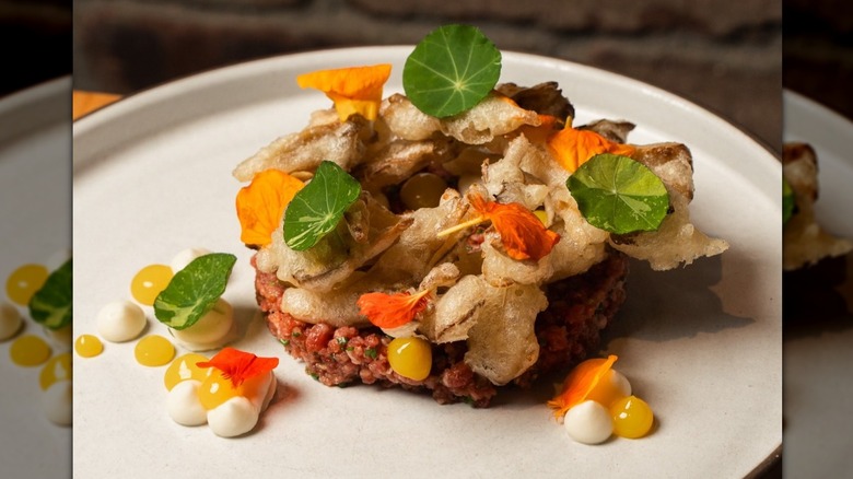 Beef tartare on white plate topped with crispy mushrooms, edible flowers, green leaf flourishes, and dollops of sauce