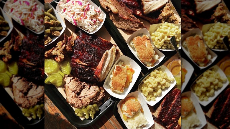 Huge array of barbecued meat with crispy bark, assorted sides, and garnishes on wood table, including sliced brisket, chicken, coleslaw, mac and cheese, pickles, and more