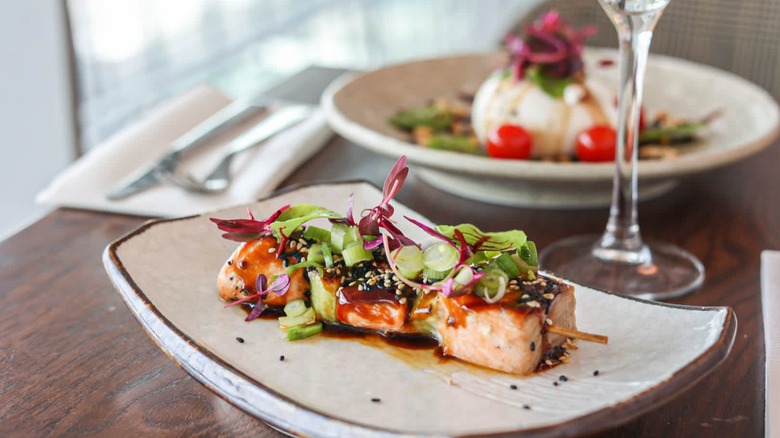 Skewer with greens on a white plate with a wine glass and another dish blurred in the background