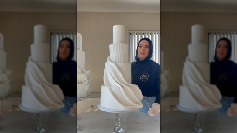 Person standing next to a white, four-tier wedding cake with a fondant drape