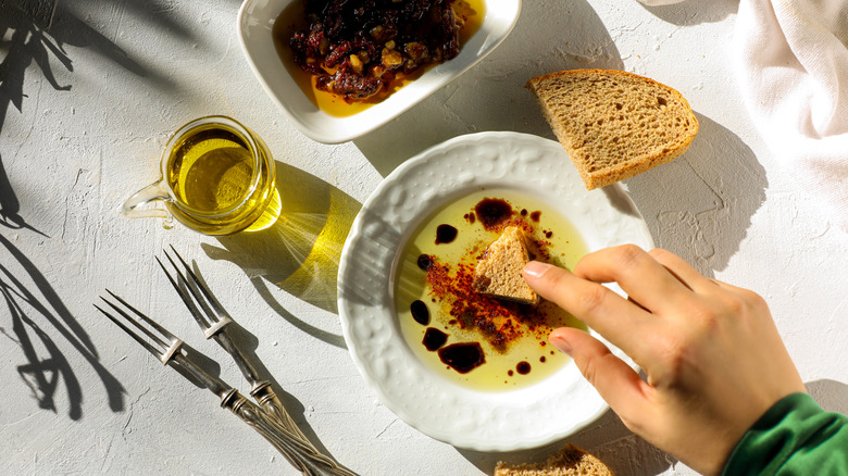 A person's hand is pictured dipping a brown slice of bread into a dish containing olive oil and balsamic vinegar.