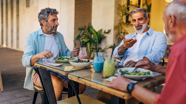 Three older men enjoy salads and colorful drinks together at an outdoor table at a Spanish restaurant.