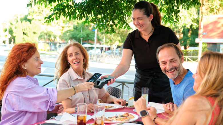 Friends dining at an outdoor restaurant pay a waitress by tapping a credit card on a device.