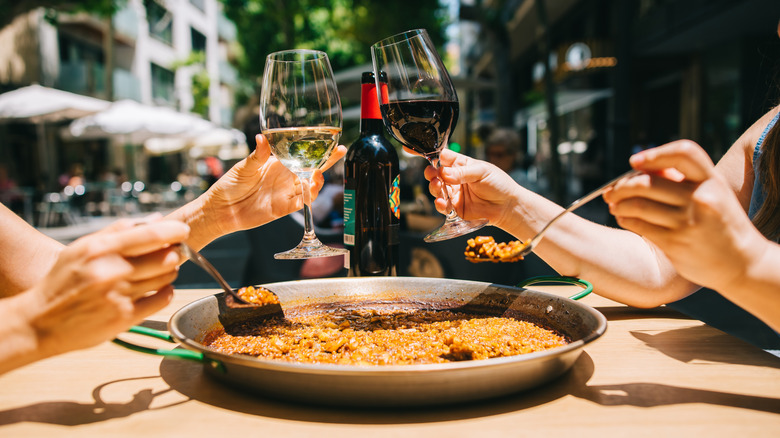 Two individuals toast their wine glasses while simultaneously digging into a plate of seafood paella in Spain.