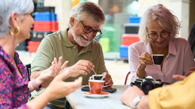Three older adults smile while enjoying small cups of coffee at an outdoor cafe in Spain.