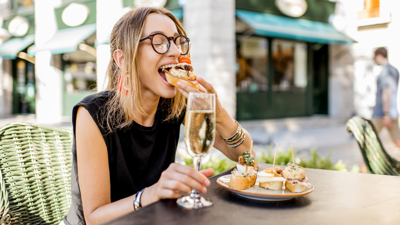 A woman takes a bite of pinchos (or "pintxos") in Spain while holding a glass of wine as she sits at an outdoor table.