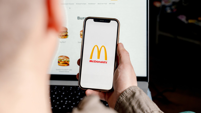 A person looking at their phone, which shows the McDonald's logo, with a menu shown on a laptop in the background