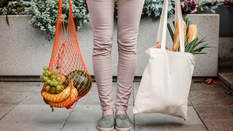 A woman holds a red mesh produce bag and a white canvas bag with groceries inside