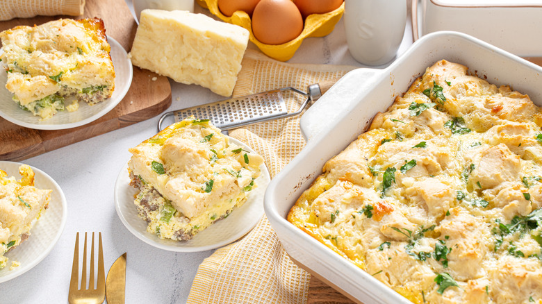 Breakfast casserole with biscuits in a dish, with slices on two plates next to eggs, a cheese grater and a hunk of cheese