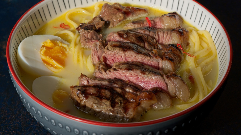 a bowl of ramen with cloudy broth topped with sliced steak and hard-boiled eggs
