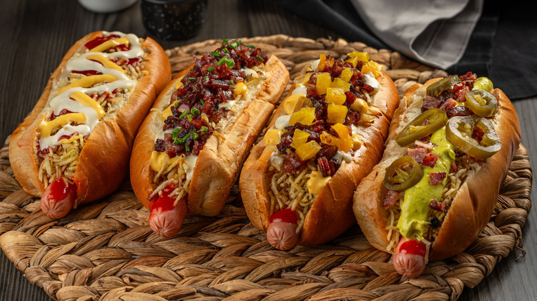 Four hot dogs with a variety of toppings including chili, pickled jalapenos, cheese, mustard, mayonnaise, and guacamole