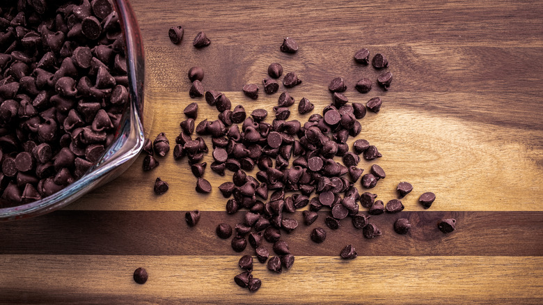 Bowl of chocolate chips next to some loose chocolate chips on a wooden cutting board