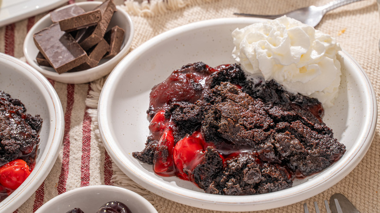 A bowl of rich, gooey, chocolate dump cake with cherries and whipped cream next to a bowl of chopped chocolate