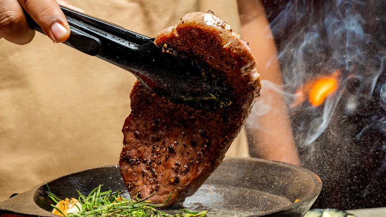 A hand flips a pork steak in a sizzling skillet