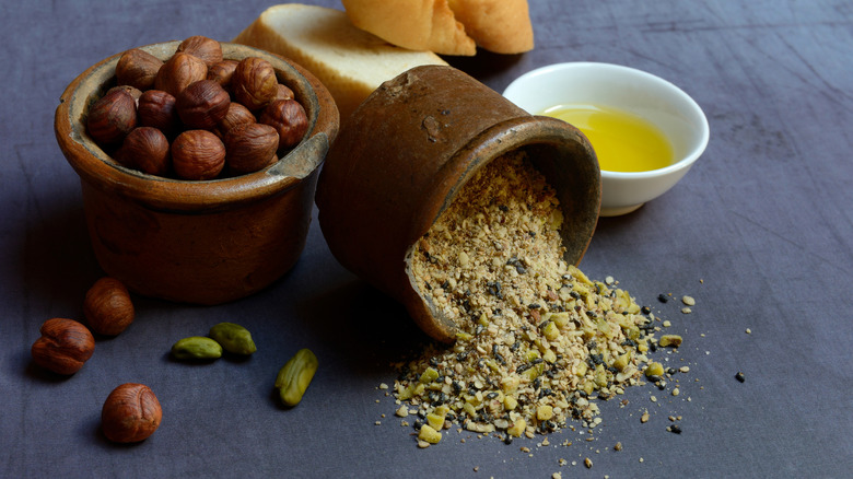 Dukkah spilling out of a ceramic bowl surrounded by hazelnuts, pistachios, bread, and olive oil