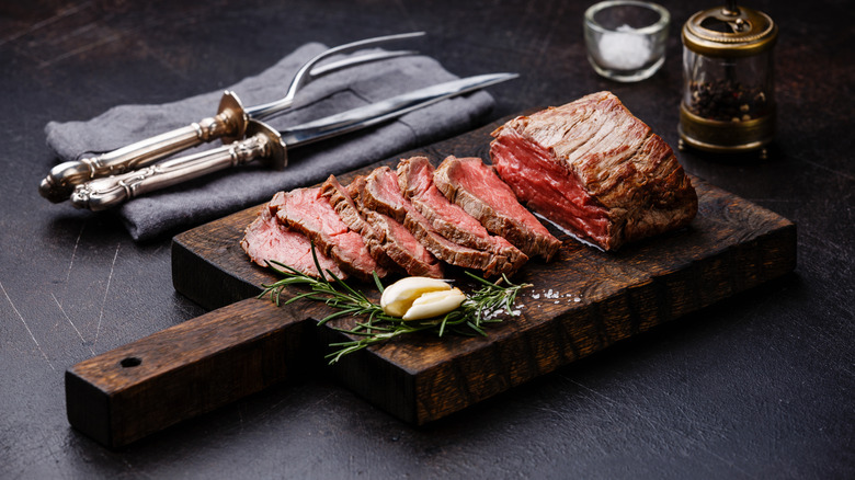 A peice of roast beef and slices on a wooden cutting board with rosemary, garlic, salt, and pepper, resting alongside serving cutlery on a grey napkin.