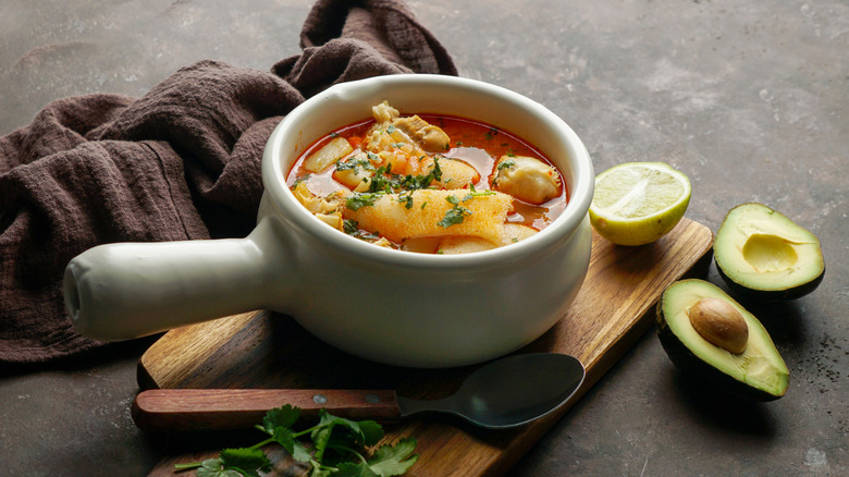 Mondongo soup in a white bowl on a wooden board, next to a halved lime and avocado