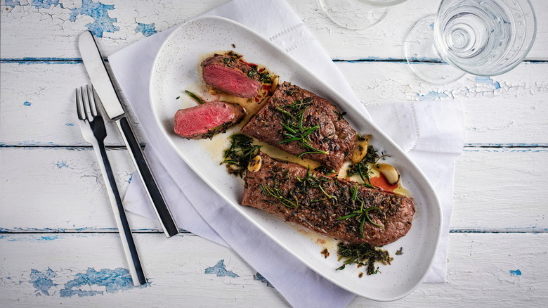 A plate of grilled venison backstrap on a white wooden table with cutlery and a glass