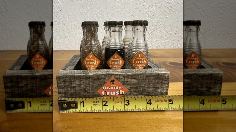 wooden crate of miniature Orange Crush Bottles against white wall with measuring tape