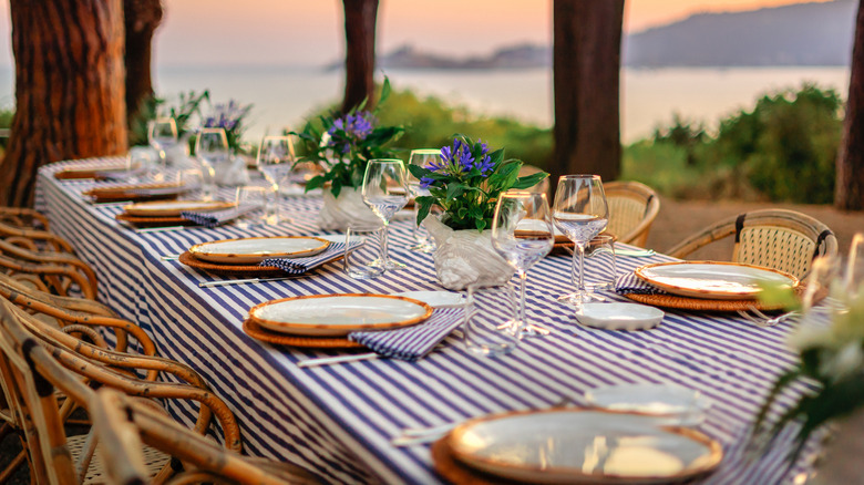 A table with a blue and white cloth set up with dishware, overlooking trees and the ocean
