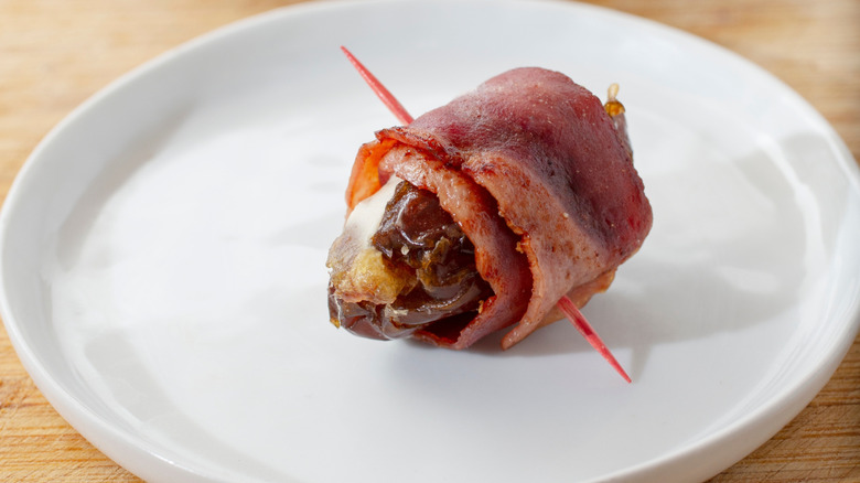 Bacon-wrapped prune with a toothpick on a white plate