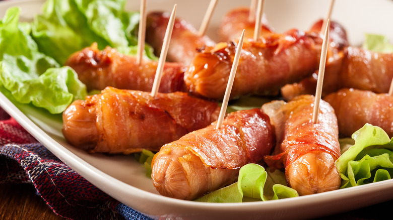 Mini sausages wrapped in smoked bacon on a plate with lettuce and toothpicks