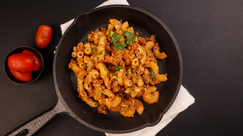 Slumgullion in a black skillet on a black table next to a dish of tomatoes