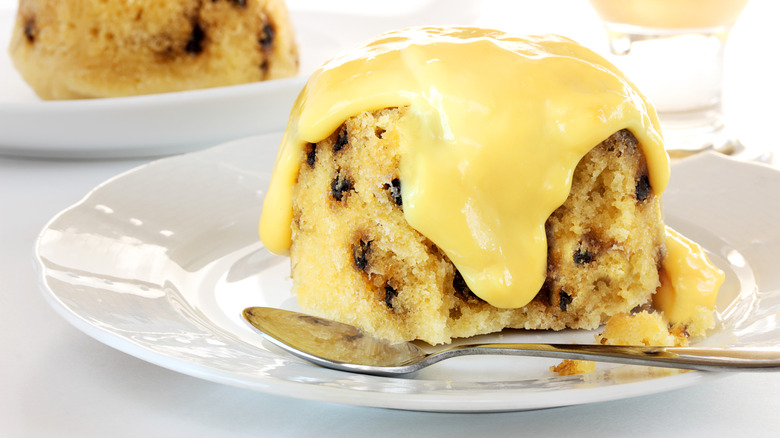 Spotted dick on a white plate with a spoon and lots of custard on top