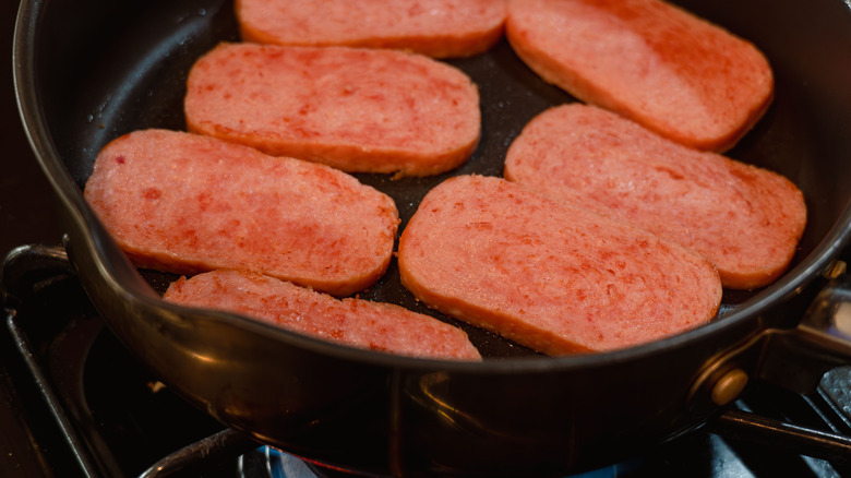 Slices of Spam frying in a black pan over a gas stove with a blue flame