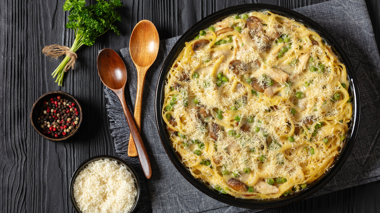 Turkey tetrazzini in black casserole dish, next to bowls of shredded cheese and peppercorns