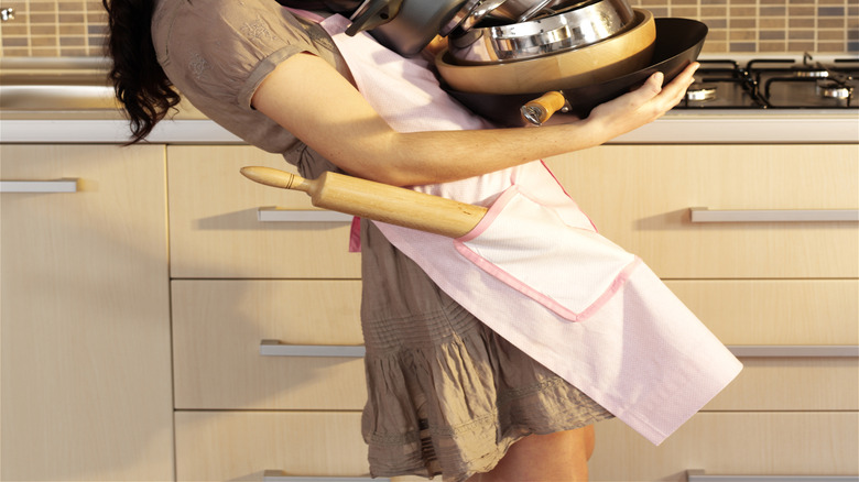 Laughing person carrying an armful of pots and pans in a kitchen