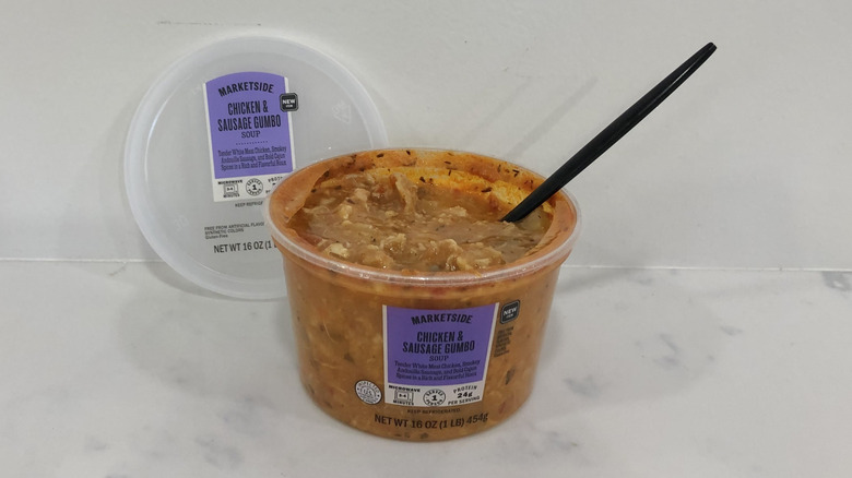 Opened plastic tub of Walmart Marketside Chicken & Sausage Gumbo with black spoon on white surface